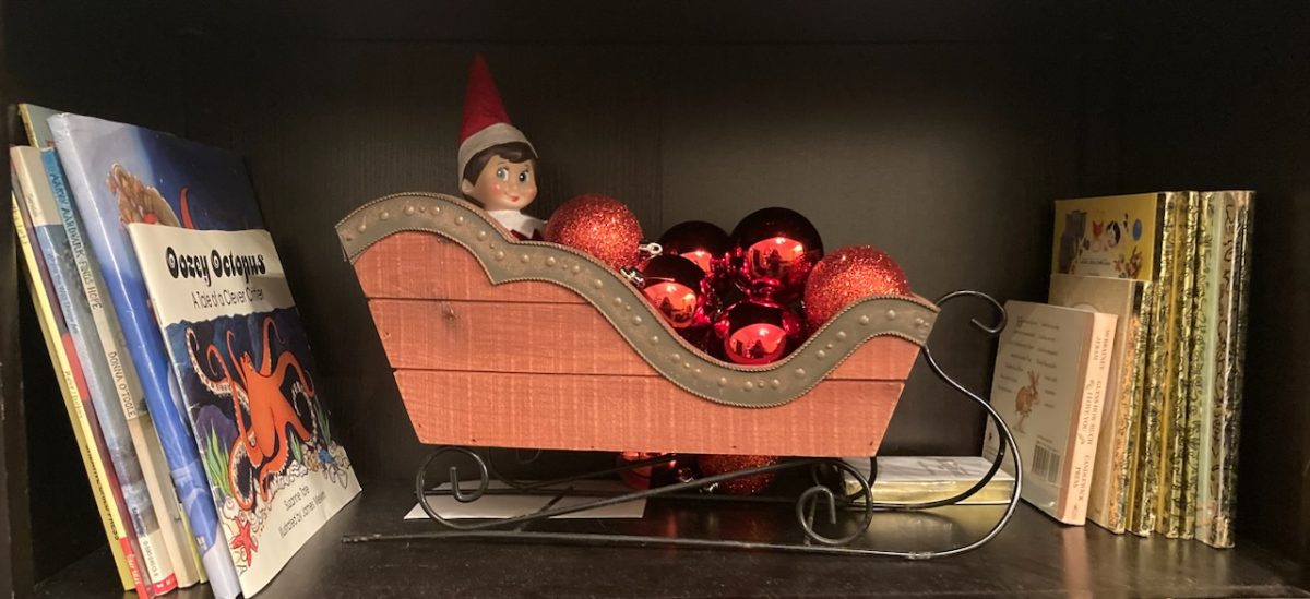 The Elf of the Shelf Twinkle sits in a miniature sleigh engulfed in Christmas ornaments. 