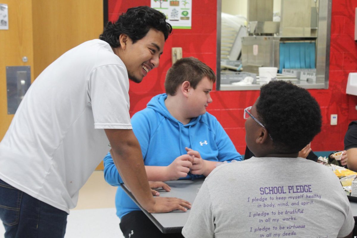 Senior Jayfren Ortigoza stands talking and laughing with a table of eighth grade boys. 